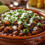 A rustic clay bowl filled with creamy Anasazi beans and their deep-flavored pot liquor, garnished with fresh cilantro, crumbled white cotija cheese, a lime wedge, and served with a side of golden cornbread.